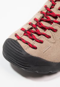 Beige suede shoe with black rubber sole. Features red and black patterned laces, textured surface, and subtle stitching details.