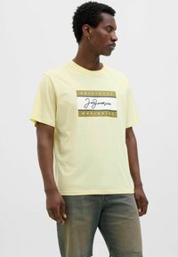 Gul bomulds T-shirt med korte ærmer, prydet med en frontgrafik: "ORIGINALS" og "WORLDWIDE" i sort og guld, med et signaturdesign.