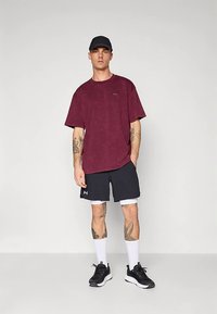 Bordové tričko z bavlny s krátkymi rukávmi, kombinované s čiernymi šortkami. Čierne športové topánky, biele ponožky a čierna čiapka. Jemný detail loga.