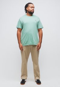 Άνδρας με πλεγμένα μαλλιά που φοράει πράσινη μπλούζα μέντας, μπεζ παντελόνι και μαύρα αθλητικά παπούτσια, στέκεται με τα χέρια χαλαρά δίπλα στο σώμα.