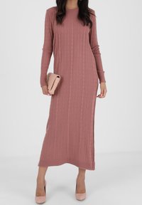 Nainen, joka pukeutuu pitkään mauven väriseen lantiossa neulottuun mekkoon, pitää kädessään vaaleanpunaista clutch-laukkua ja on pukeutunut vaaleanpunaiseen korkokenkään valkoista taustaa vasten.