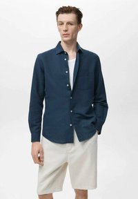 Námornícky modrá ľanová košeľa s dlhými rukávmi, so zapínaním na gombíky, s jednou náprsnej kapsou, nosená cez biele tielko, v kombinácii s svetlými šortkami.