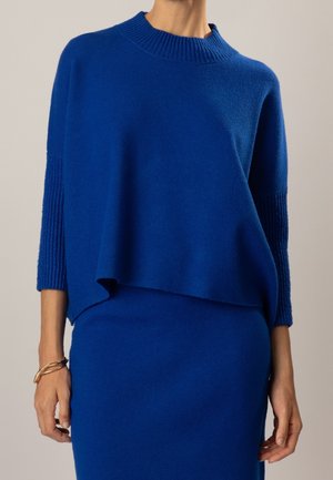Femme portant un pull en maille bleu cobalt avec des manches côtelées et une jupe bleue assortie sur un fond uni.