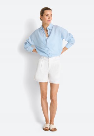 Donna con camicia azzurro chiaro con bottoni, pantaloncini bianchi e sandali bianchi intrecciati slip-on, in piedi con le mani sui fianchi, guarda di lato.