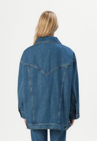 Oversized denim jacket in medium blue. Features wide collar, back yoke detailing, and button accents on sleeves. Smooth texture.