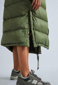 Olivza zaļa pūkainā jaka ar kvadrāto šuvēm un spiedpogu aizdari, kombinēta ar pelēkiem New Balance sporta apaviem, kuriem ir šņores un acs akcenti.