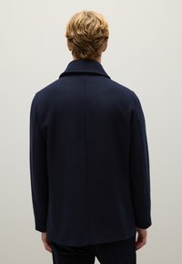 Cappotto in lana blu navy con colletto strutturato, maniche lunghe e taglio dritto, caratterizzato da un design pulito e minimalista senza chiusure visibili.