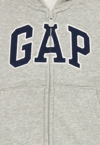 Felpa grigia con zip, caratterizzata da un grande logo "GAP" blu navy, tessuto morbido, due tasche frontali e cappuccio standard.