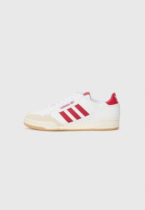 White athletic shoes with red accents and three stripes design. Features a suede toe cap and rubber sole. Minimalist design and texture.