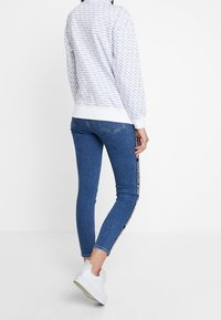 Felpa bianca con motivo a logo ripetuto, abbinata a jeans blu con striscia logo laterale e sneaker bianche.