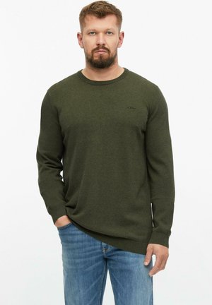 Man with beard wearing an olive green sweater and blue jeans, standing with one hand in pocket against a white background.