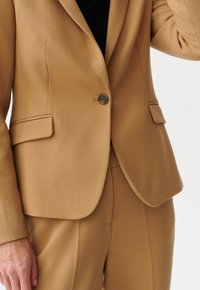 Blazer in kameelkleur met een enkele knoopsluiting, twee steekzakken aan de voorkant en een gestructureerde stof, gecombineerd met bijpassende op maat gemaakte broek.