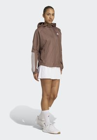 Braune leichte Jacke mit Kapuze, die weiße Drei-Streifen-Akzente an den Ärmeln aufweist. Getragen über einem weißen Rock und kombiniert mit weißen Sneakers.