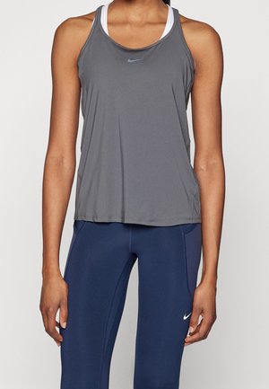 Atletyczna osoba ubrana w szary top Nike na biały sportowy stanik i granatowe legginsy Nike, stojąca z rozluźnionymi ramionami.