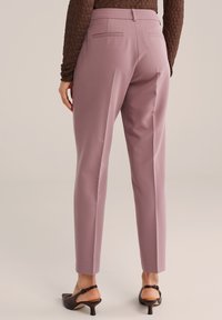 Spodnie na miarę z wysokim stanem, uszyte z matowej tkaniny w kolorze przygaszonego mauve. Posiadają dwie tylne kieszenie, zakładkę z przodu oraz zwężane nogawki. Noszone z sandałami na obcasie.