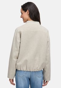 Beige kurz geschnittene Jacke mit Kragen, texturiertem Stoff und gerafftem Saum. Getragen über hellblauen Jeans, von hinten betrachtet.