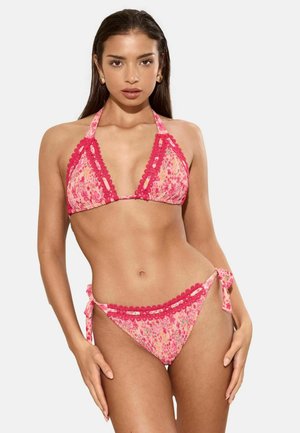 Woman wearing a pink floral two-piece bikini with red lace trim and side ties, standing against a plain white background.