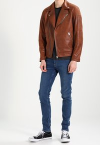 Giacca da moto in pelle marrone con dettagli a coste sulle spalle, due tasche con zip, abbinata a jeans scuri e sneaker nere.