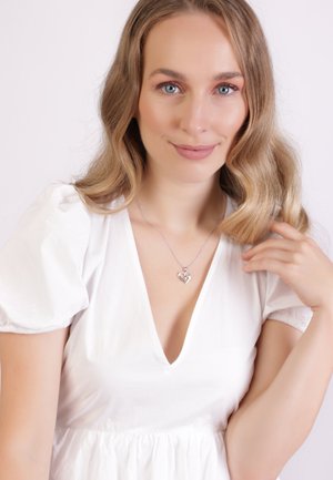 Zilveren hart hanger ketting met een heldere steen, gedragen door een model met lang, golvend haar, gekleed in een witte jurk met pofmouwen.