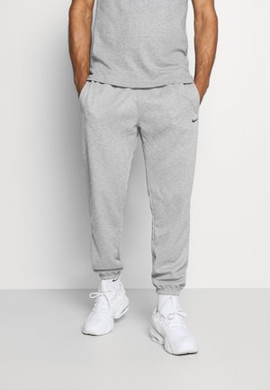 Mężczyzna ubrany w jasnoszare spodnie dresowe Nike, pasującą koszulkę, białe sneakersy Nike oraz białe skarpetki, stojący na białym tle, z rękami w kieszeniach.