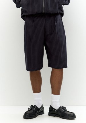 Shorts à fines rayures bleu marine avec une coupe décontractée, dotés de poches latérales et d'une ceinture. Associés à des chaussures noires et des chaussettes blanches.