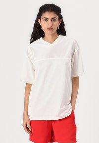Haut court à manches courtes en mesh blanc avec un col en V, comportant une section supérieure unie et un tissu perforé en dessous. Associé à un short rouge.