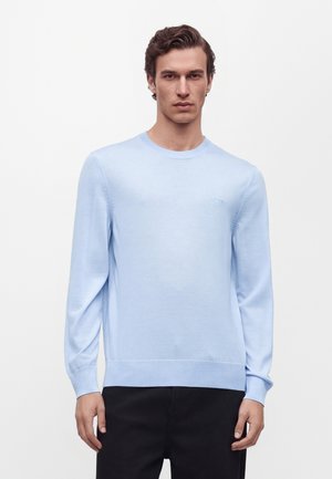 Homme aux cheveux courts et bruns portant un pull bleu clair à manches longues et un pantalon noir sur un fond blanc uni.
