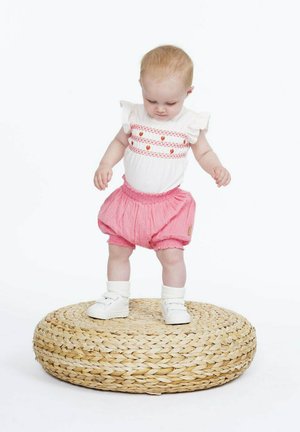 Haut en coton blanc avec broderies rouges, associé à un short bouffant rose texturé. L'enfant se tient sur un tapis rond en paille tissée en portant des chaussures blanches.
