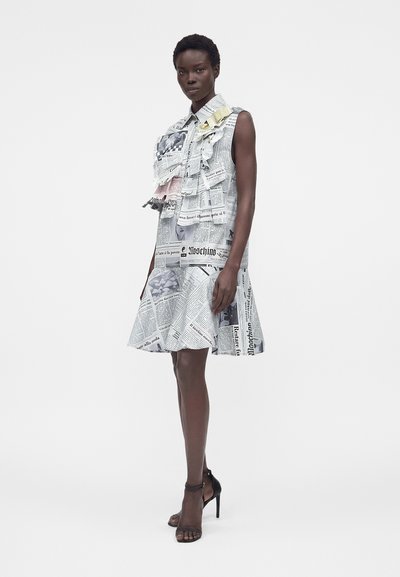 Μαύρη γυναίκα που φοράει αμάνικο φόρεμα με τύπωμα εφημερίδας και διακοσμητικά βολάν, στέκεται με το ένα χέρι χαλαρό, φοράει μαύρα ψηλοτάκουνα σανδάλια.