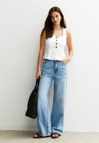 Haut sans manches blanc avec des boutons ronds, associé à un jean large bleu clair. Un sac tissé noir complète la tenue.