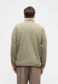 Άνδρας που φοράει μπεζ υφασμάτινο τζάκετ φλις με υφή και καφέ παντελόνι, στέκεται με την πλάτη στην κάμερα μπροστά σε απλό λευκό φόντο.