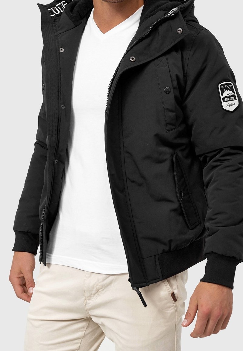 Zwarte zip-up jas met een gevoerde capuchon, zijzakken en een patch op de linker mouw. Gepaard met een witte v-hals t-shirt.