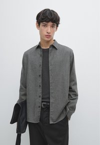 Camicia grigia con bottoni caratterizzata da una texture morbida, colletto classico e maniche lunghe, abbinata a una cintura e pantaloni neri. Giacca nera in mano.