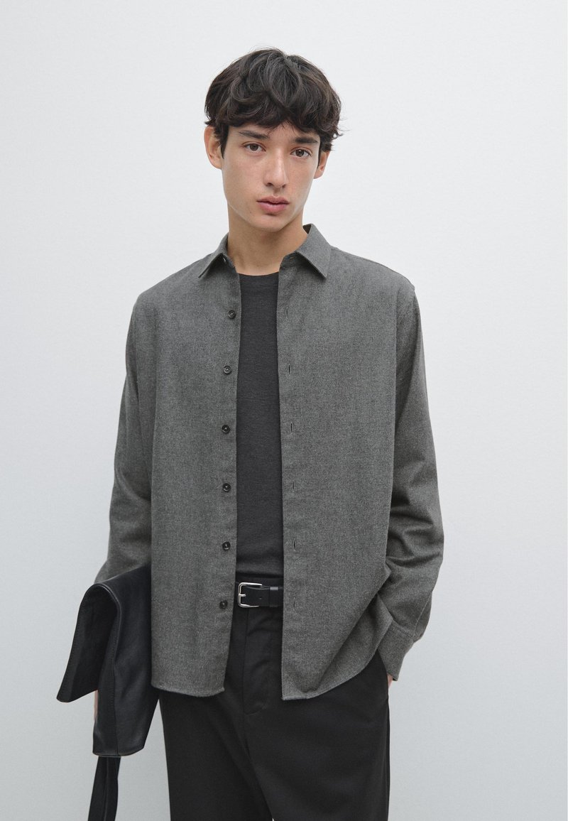 Camicia grigia con bottoni caratterizzata da una texture morbida, colletto classico e maniche lunghe, abbinata a una cintura e pantaloni neri. Giacca nera in mano.