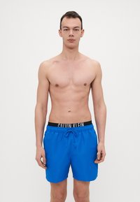 Blaue Badehose mit Kordelzug, ausgestattet mit einem schwarzen Bund, der mit "CALVIN KLEIN" beschriftet ist. Glatte Textur, Länge bis über das Knie.