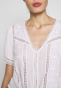 Weiße Baumwollbluse mit aufwendiger floraler Bestickung, Spitzenbesatz und tiefem V-Ausschnitt. Puffkurze Ärmel mit Ausschnittdetails.
