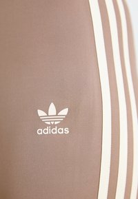 Rudos sportiniai legginsai su trimis baltomis juostelėmis šonuose ir balta Adidas logotipu atspausdintu prie apačios. Lygus audinio paviršius.
