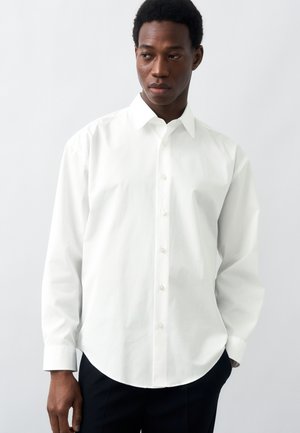 Homme portant une chemise blanche unie à manches longues boutonnée et un pantalon noir, regardant sur le côté sur un fond blanc.