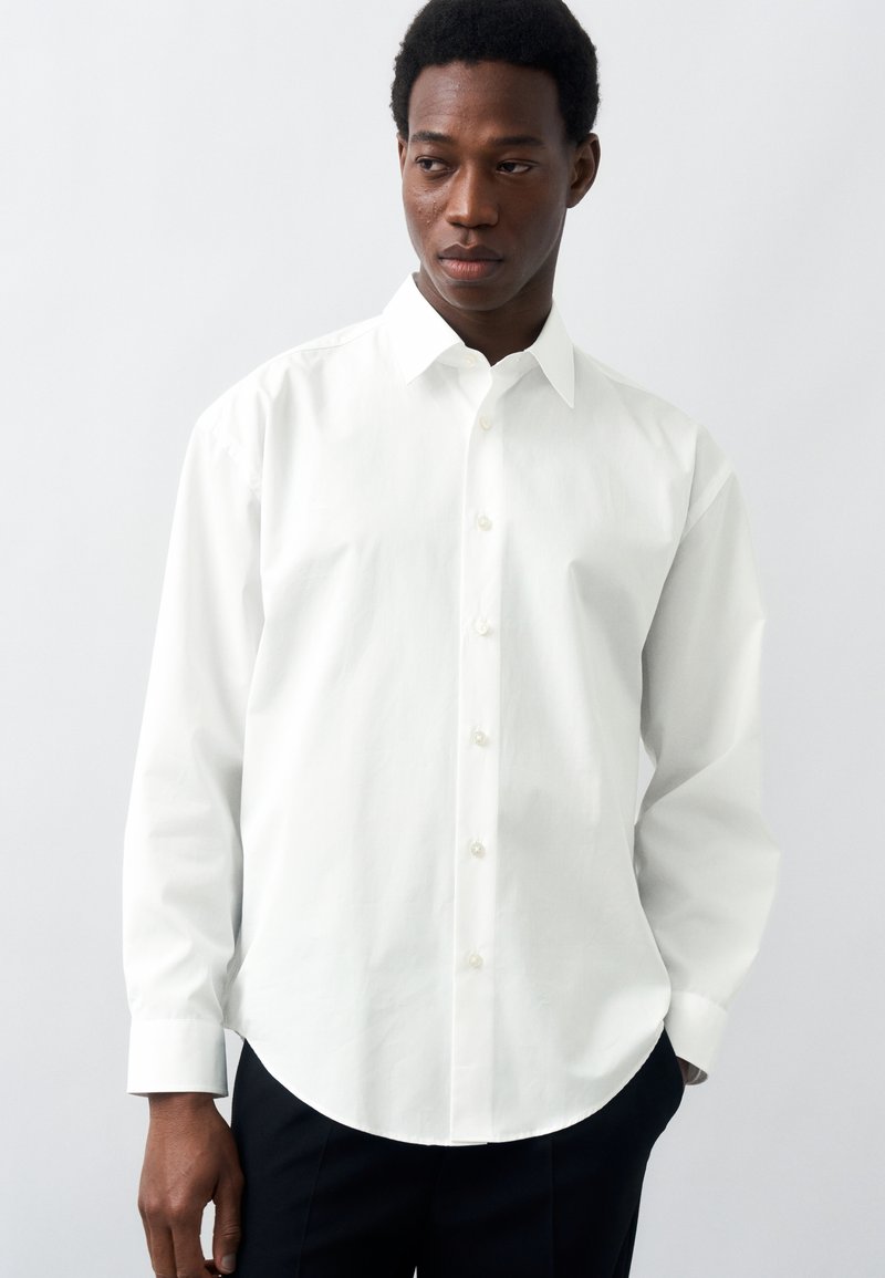 Homme portant une chemise blanche unie à manches longues boutonnée et un pantalon noir, regardant sur le côté sur un fond blanc.