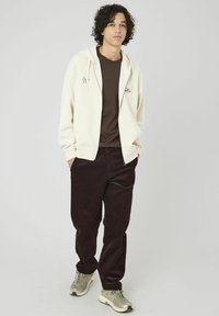 Haut à capuche crème avec cordons, t-shirt marron en dessous, pantalon en velours côtelé sombre et baskets grises avec des accents en mesh. Tenue simple et décontractée.