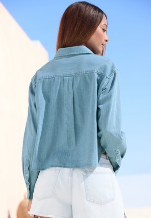 Jonge vrouw met recht, bruin haar, gekleed in een lichtblauw denim shirt en witte korte broek, staande buiten tegen een heldere blauwe lucht.