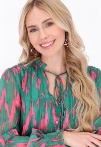 Blusa colorata con un motivo astratto verde e rosa, scollatura arricciata con lacci e polsini arricciati. Tessuto liscio e leggero.