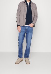 Outfit casual con giacca a quadri beige sopra una maglietta blu con colletto a zip, jeans blu e scarpe in suede taupe. Design semplice e contemporaneo.