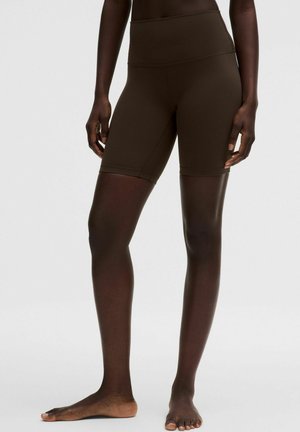 Sötétbarna testhezálló motoros rövidnadrágot viselő személy áll mezítláb egy egyszínű világos háttér előtt.