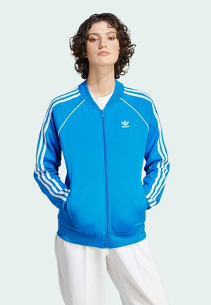 Blå atletisk zip-up jakke med hvide tre-stribede detaljer på ærmerne og broderet logo på brystet. Har lommer og ribbede manchetter.