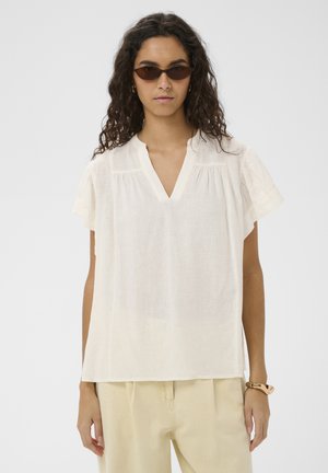 Vrouw met krullend haar, donkere zonnebril, een losse witte blouse met korte mouwen en V-hals, beige broek en een dikke armband om haar pols.