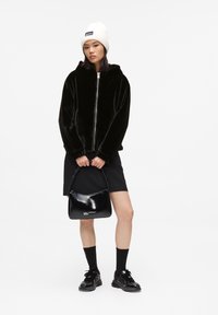 Black faux fur zip-up hoodie with a hood, paired with a black mini skirt. Holding a glossy black handbag, black socks, and sporty black shoes.