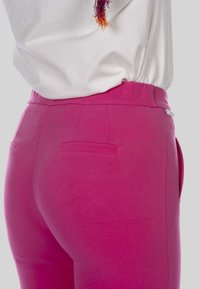 Rooze stretchbroeken met een gladde textuur, voorzien van achterste paspelzakken en een platte elastische tailleband. Een accentlogo is zichtbaar aan de zijkant.