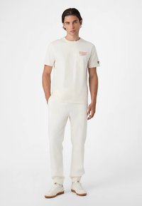 Beige puuvillainen pyöreäkauluksinen t-paita, jossa lyhyet hihat ja punainen logotunnus, yhdistetty samansävyisiin collegehousuihin. Asu tarjoaa rentoutuneen istuvuuden.