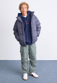 Giacca a vento grigia con cappuccio, indossata sopra un felpa blu navy con un logo rosso, abbinata a pantaloni cargo verdi e sneakers bianche.